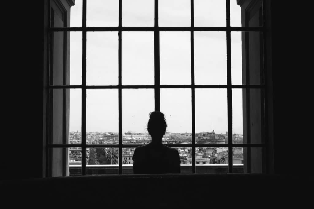 A black and white shot of a lonely female standing in front of the windows looking at the buildings